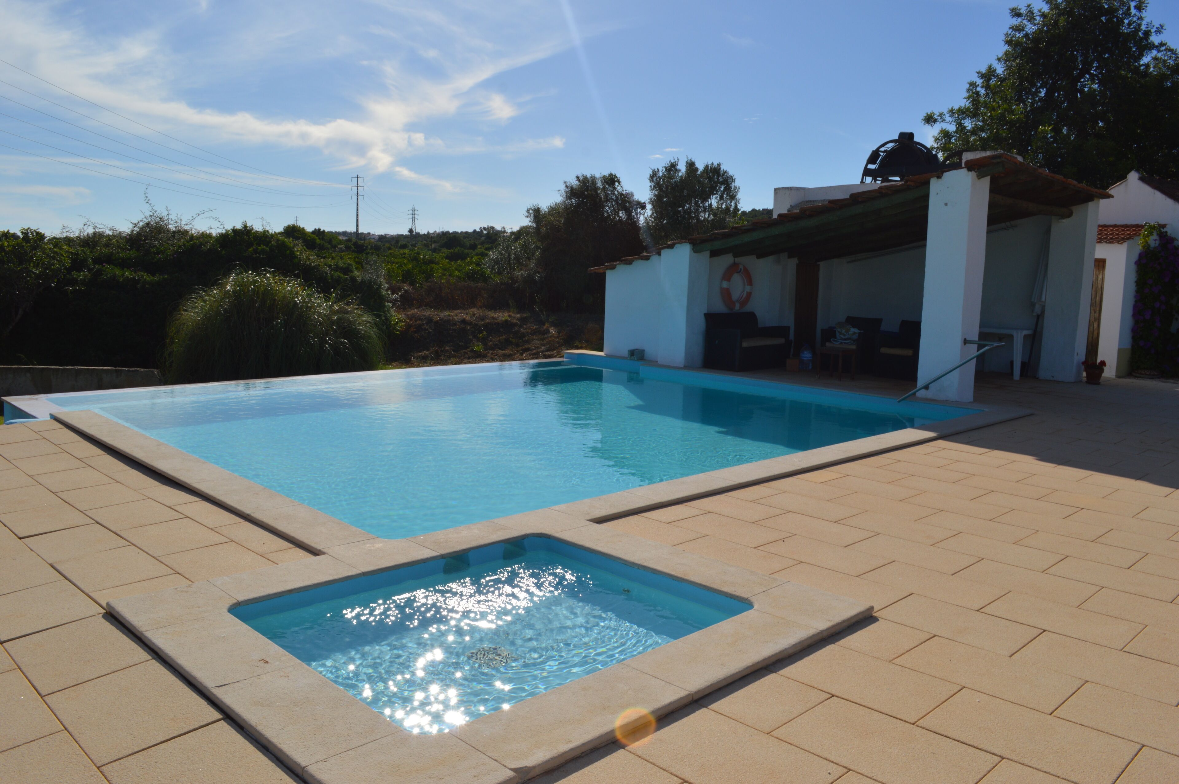 Piscina con zona a livello ribassato, circondata da un solarium piastrellato e un’area relax coperta, con un salvagente in vista.