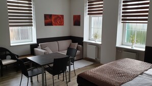 A modern room with a sofa, a dining table, and chairs, a bed with a patterned blanket, and two windows with blinds. - Apartma Transpet (Kostelec na Hané)