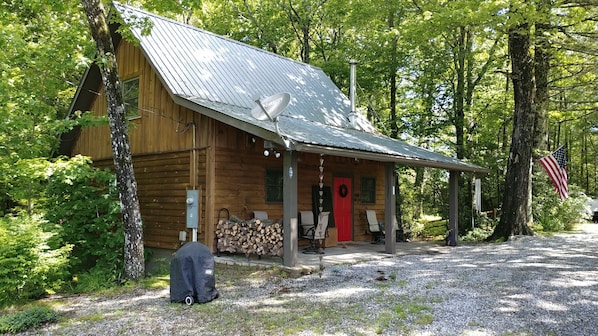 A wooden cabin with a metal roof, a porch with chairs, and a stack of firewood.