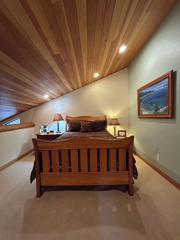 A bedroom with a wooden ceiling, a bed with a headboard, two bedside tables, a lamp, and a framed picture on the wall.