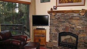 Una sala de estar con chimenea de piedra, muebles de cuero, un televisor sobre un soporte de madera y un ventanal con vistas a árboles.