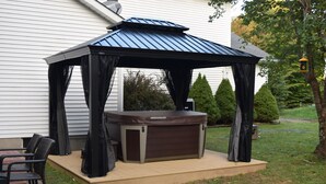 A hot tub with a canopy, surrounded by chairs, in a backyard with a house and trees.