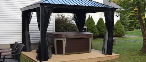 Un jacuzzi con toldo, rodeado de sillas, en un patio trasero con una casa y árboles.
