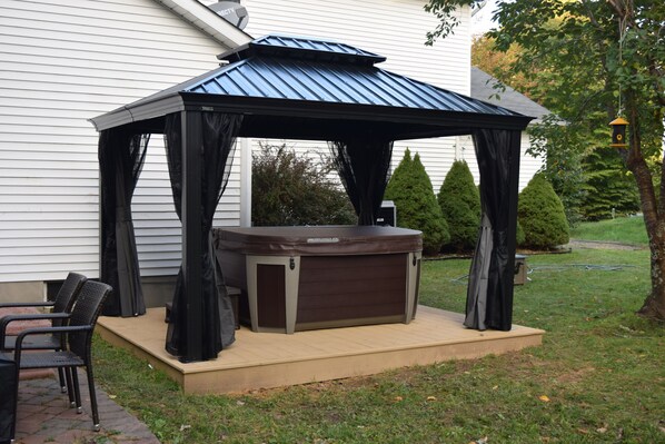 A hot tub with a canopy, surrounded by chairs, in a backyard with a house and trees.