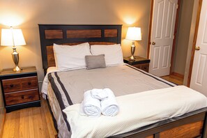 A neatly made bed with pillows and a folded towel, flanked by bedside tables with lamps, in a room with wooden flooring and a door in the background.