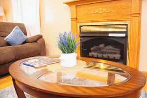 A living room with a brown sofa, a glass coffee table with a white vase of flowers, and a fireplace with a log fire. - Luxurious Villa in Pocono near Ski Slopes with Game room & Hot tub & Golf (Tobyhanna)