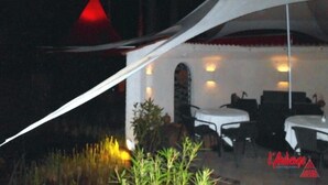 Un área de comedor al aire libre con mesas y sillas blancas, una carpa blanca y una luz roja al fondo.