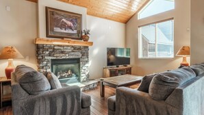 A cozy living room with a stone fireplace, a flat-screen TV, and a large window with blinds. - Mammoth Gateway Village #1 - Updated Townhome-Walk To Village 3 Bed+Loft, Sleeps 10 Private Hot Tub (Mammoth Lakes)