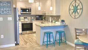 A modern kitchen with white cabinets, a central island with bar stools, a ceiling fan, and a wall-mounted compass decoration. - Gorgeous 5 Star! Oceanblock Midtown condo directly Across from Convention Center (Ocean City)