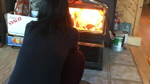 Una persona asseguda davant una estufa de llenya en una habitació amb parets de fusta i un terra de rajola.