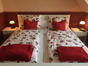 A twin bed with floral bedding and red pillows in a room with a sloped ceiling and two bedside lamps. - Friesenglück Kolkerheide (Kolkerheide)