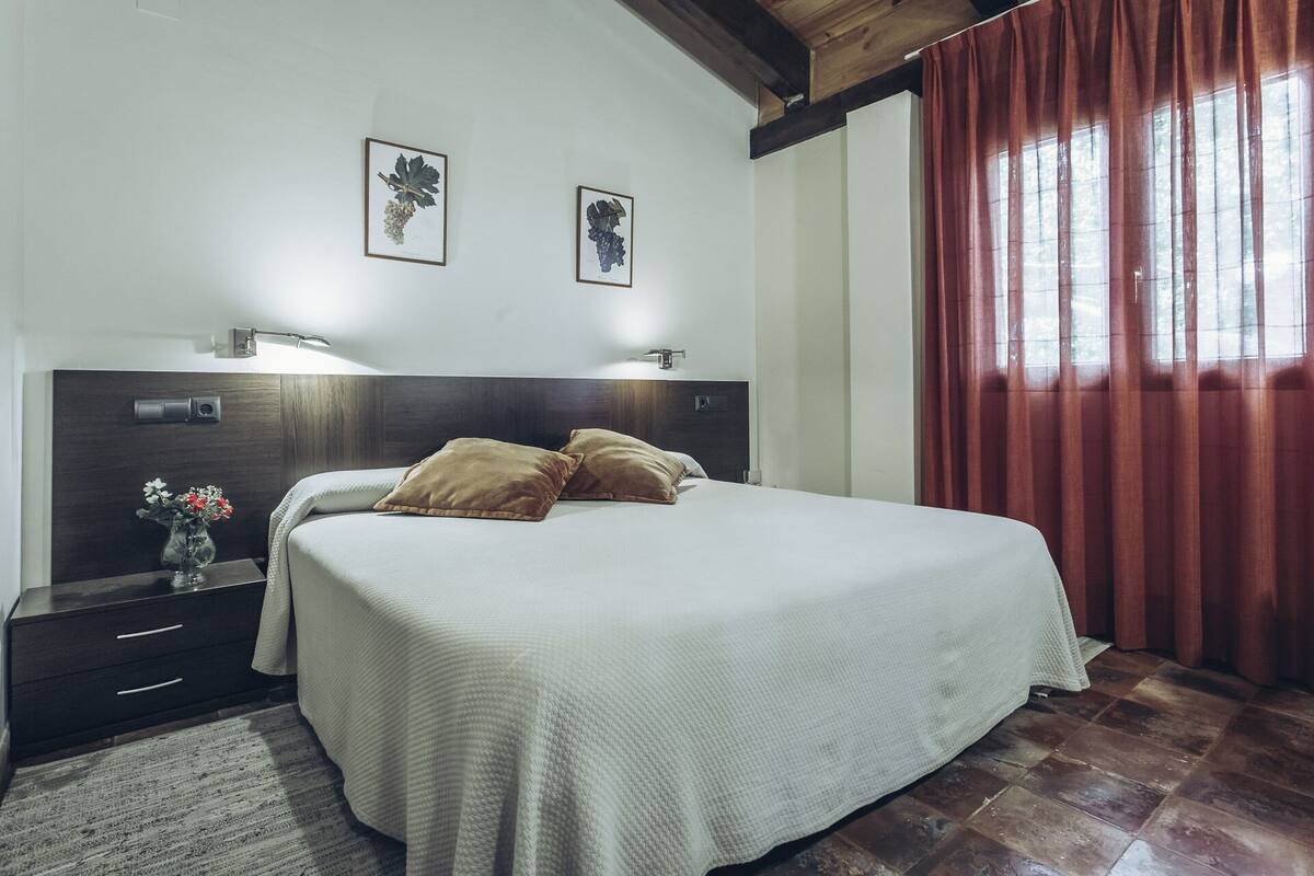 Un dormitorio con una cama, dos mesitas de noche, una lámpara de noche, un jarrón con flores y dos cuadros en la pared.
