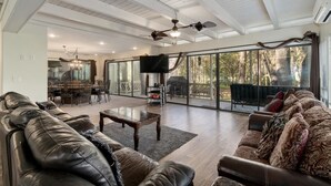Un salón amplio con un sofá grande, una mesa de centro y un televisor. Hay un comedor con una araña y un ventanal grande con vistas al exterior.
