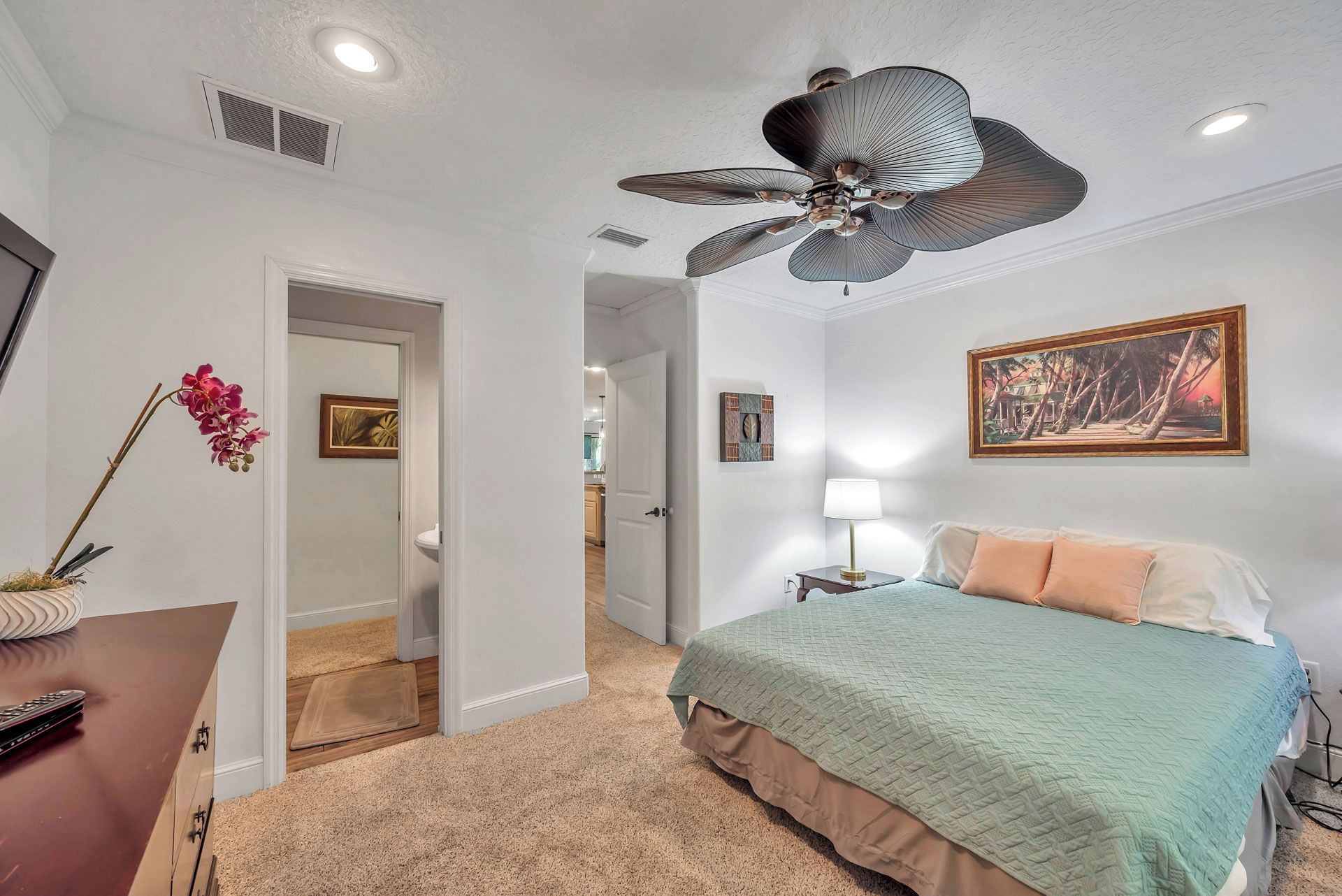 A bedroom with a bed, a desk, a chair, a ceiling fan, and a painting on the wall.