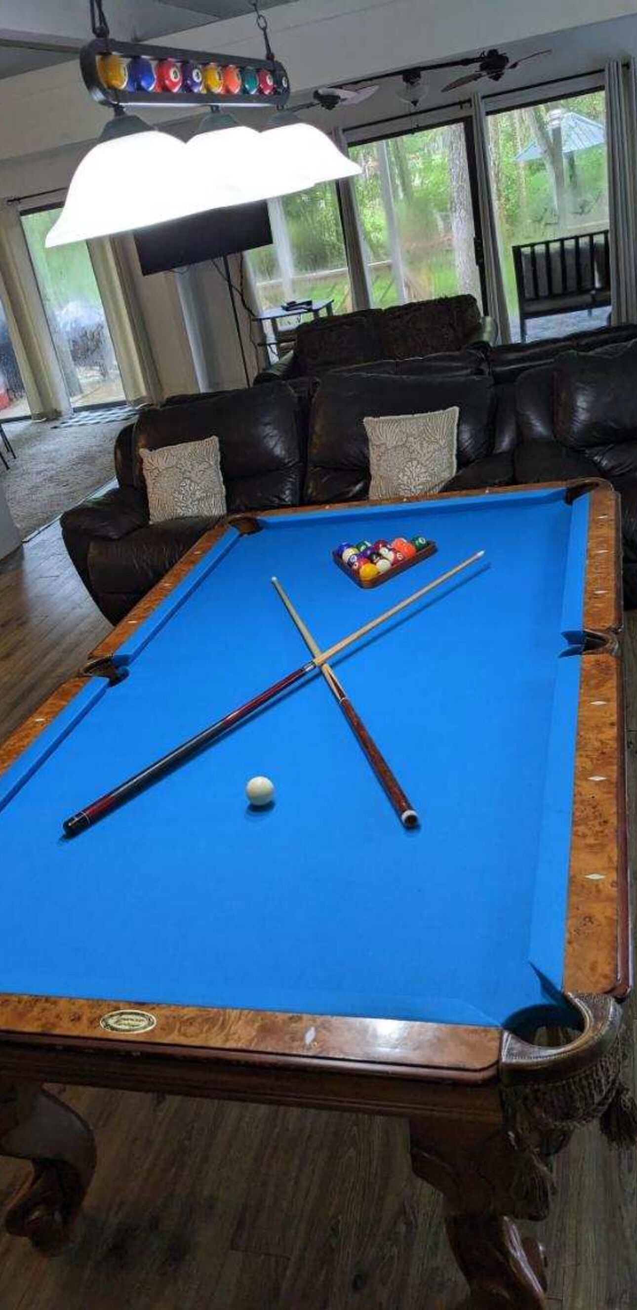 A pool table with balls and cues, a leather couch, and a large window with a view of greenery.