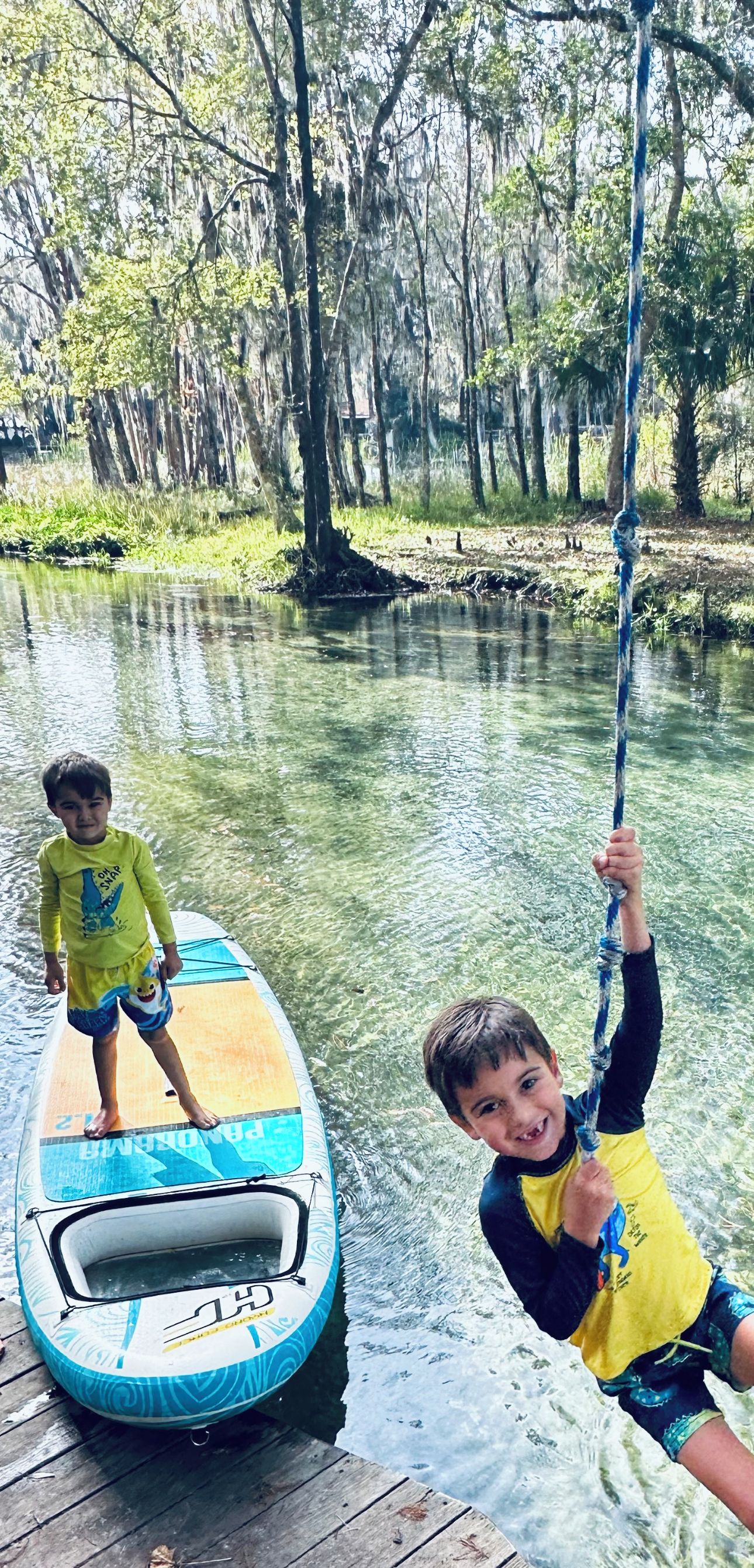 Two children on a paddleboard in a calm body of water surrounded by trees.