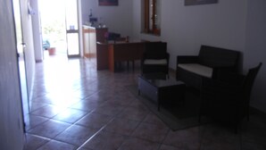 Amplia sala de estar con piso de baldosas, un escritorio de madera, un sofá y dos sillones. Hay una ventana con persianas y una puerta que da a otra habitación.