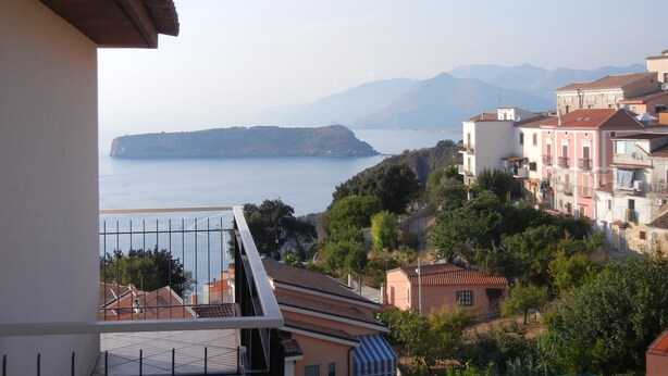 Un pueblo costero con edificios que dan al mar, un balcón con barandilla de metal y montañas al fondo.