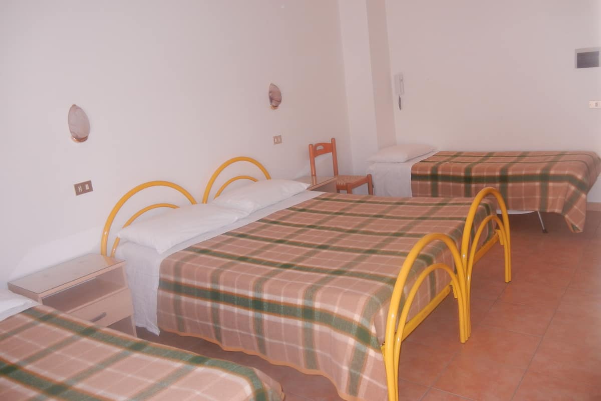 Una habitación con dos camas individuales, cada una con estructura metálica amarilla y colcha escocesa. Hay una pequeña silla de madera y una mesita de noche con una lámpara.