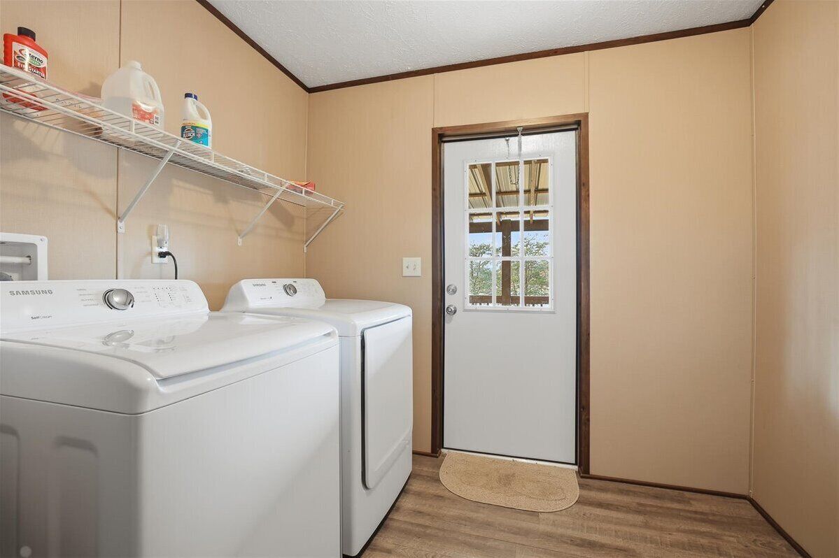 A laundry room with two white washers and dryers, a shelf with cleaning supplies, and a door with a view of greenery outside.