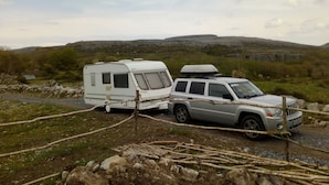 A white RV and a silver SUV parked on a gravel path with a rocky landscape and hills in the background. - Burren Bushcraft - Camping & Adventures (Carran)
