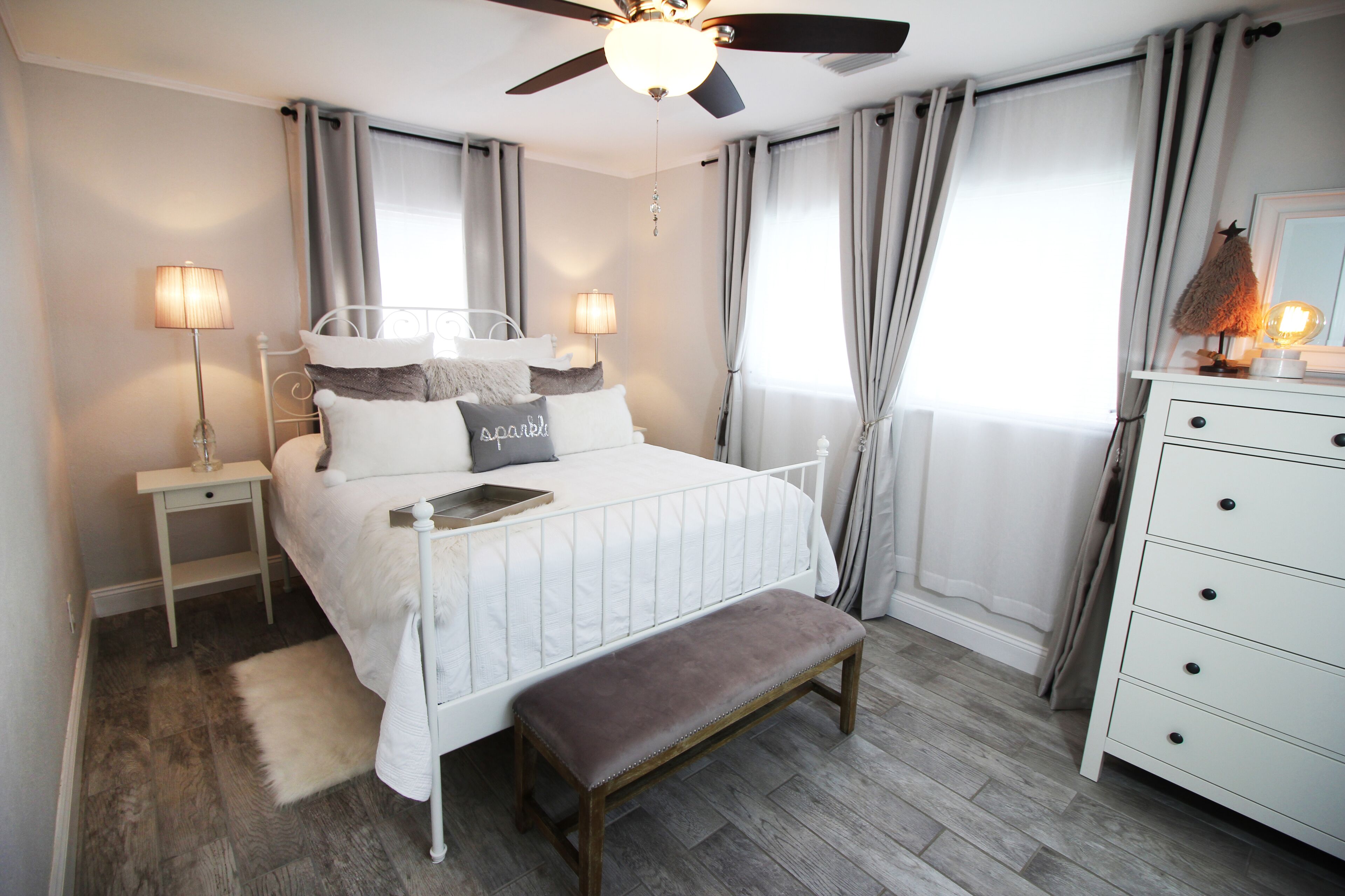 Une chambre à coucher comprenant un lit, des tables de chevet, un banc, une commode et un ventilateur de plafond.