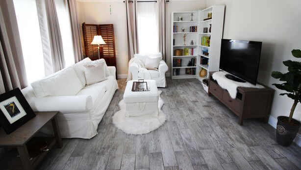 Un salon avec un canapé blanc, un fauteuil blanc, un pouf blanc, une table basse en bois, une bibliothèque, un téléviseur et un ventilateur de plafond.