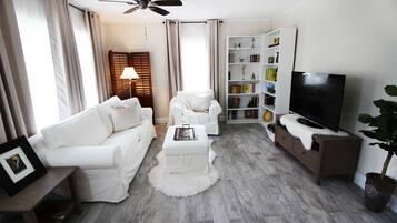 Un salon avec un canapé blanc, un fauteuil blanc, un pouf blanc, une table basse en bois, une bibliothèque, un téléviseur et un ventilateur de plafond.