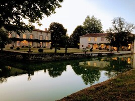 A serene lakeside view of a historic stone building with a well-maintained garden and a clear reflection in the water.