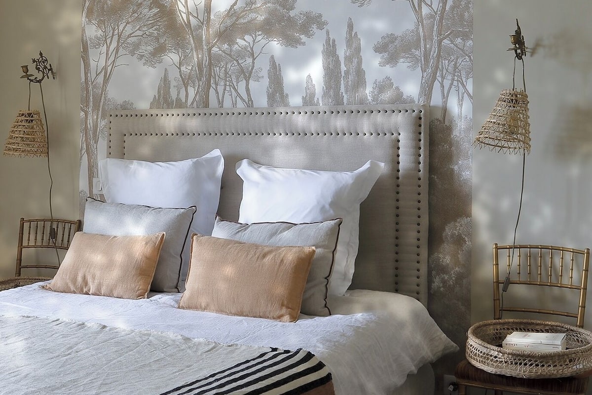 Un dormitorio con una cama grande, un cabecero con un mural de árbol, dos mesitas de noche con lámparas y una silla con cesta.