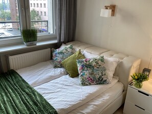A neatly made bed with floral pillows, a green throw blanket, and a potted plant on the windowsill. - Good Time Apartments Zgoda (Warsaw)