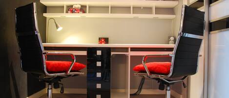 A modern office space with two black office chairs with red cushions, a desk, a lamp, and a shelving unit with various items.