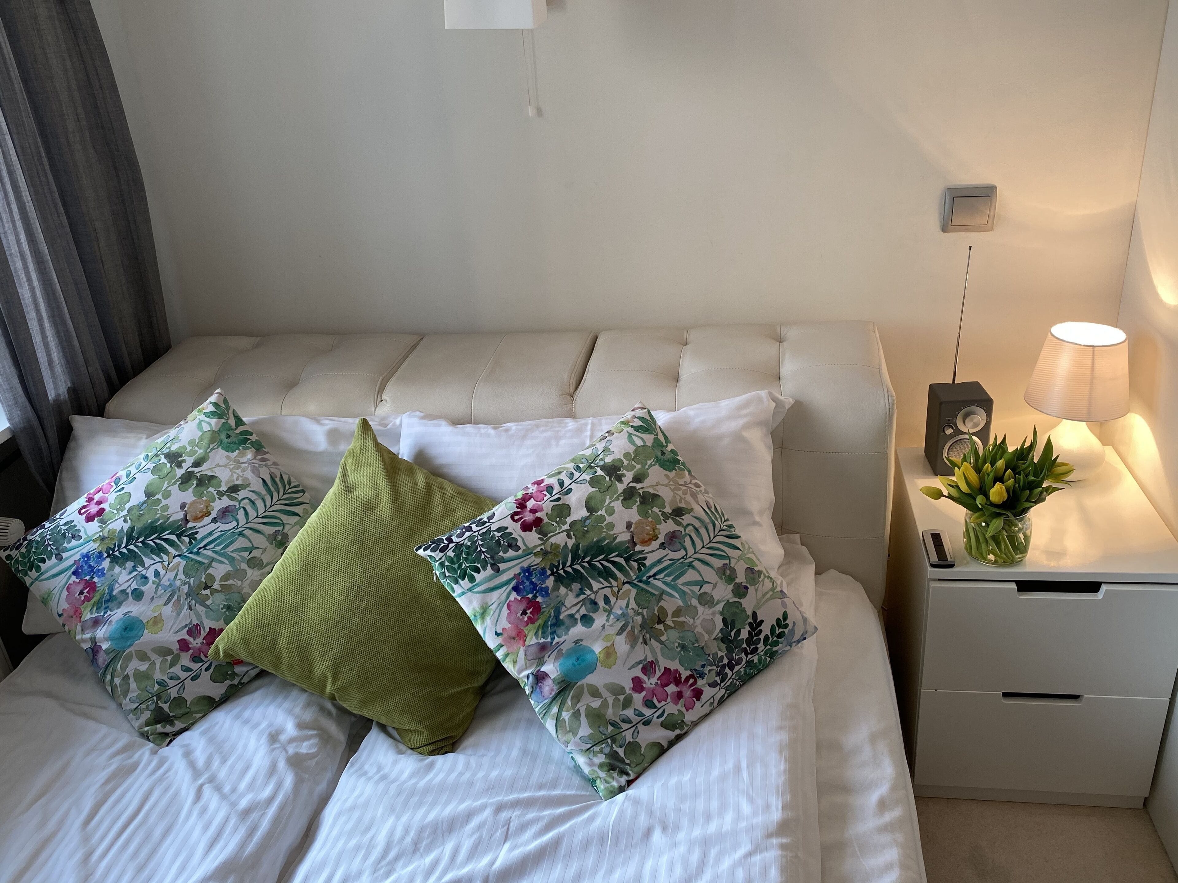 Cama bem arrumada com roupa de cama branca e almofadas estampadas com flores, uma luminária de cabeceira e uma pequena planta em vaso sobre a mesa de cabeceira.