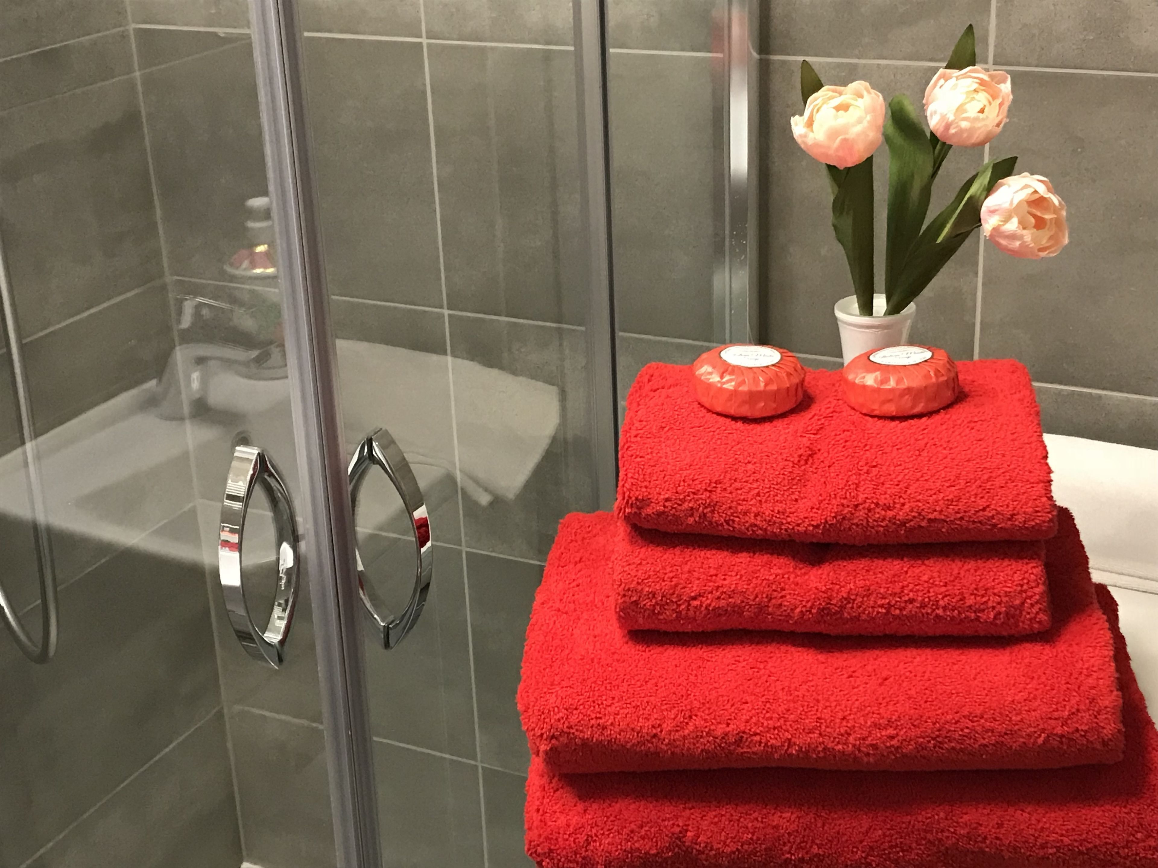 Une salle de bain avec une porte de douche en verre, une pile de serviettes rouges et un vase rempli de fleurs roses.