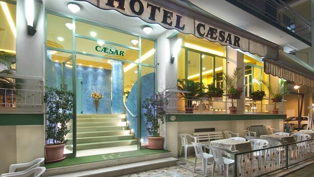 Un hotel llamado 'Hotel Caesar' con una entrada bien iluminada, zona de estar al aire libre y plantas en macetas.