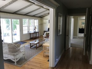 Ein geräumiges Wohnzimmer mit einem großen Fenster, Holzboden, einem weißen Korbsessel, einem Holztisch und einer Tür zu einem weiteren Raum.