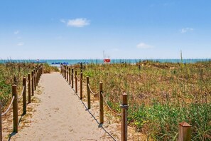 Uma trilha de areia que leva a uma praia, com uma torre de salva-vidas ao longe.