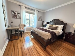 A bedroom with a wooden floor, a bed with multiple pillows, a desk with a chair, and a window with curtains.