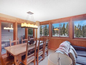 A cozy cabin interior with a dining area, a sofa, and large windows offering a view of snow-covered trees. - Nearly ski-in, walk to Gondola, one block to Main Street! (Breckenridge)