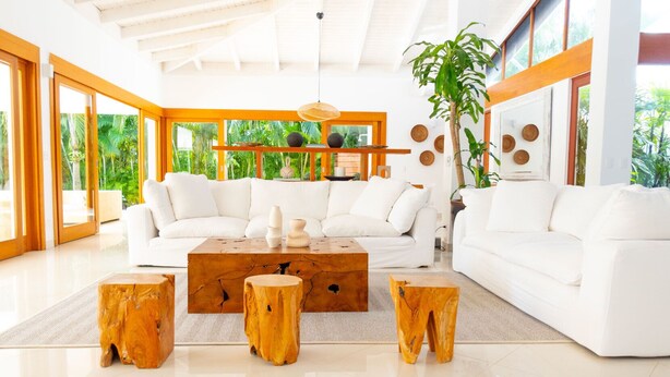 A spacious living room with a white sectional sofa, a wooden coffee table, and three wooden stools. Large windows allow natural light and views of greenery outside.