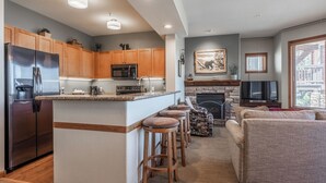 Eine moderne Küche mit Holzausstattung, einem Edelstahlkühlschrank, einer Mikrowelle und einem Steinkamin. Im Wohnbereich befinden sich drei hölzerne Barhocker und ein beiges Sofa.