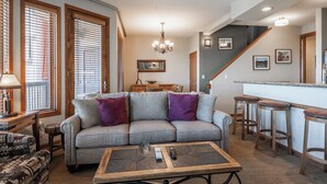 Ein Wohnzimmer mit grauem Sofa, violetten Kissen, einem Couchtisch und einer Treppe, die in einen Küchenbereich führt.