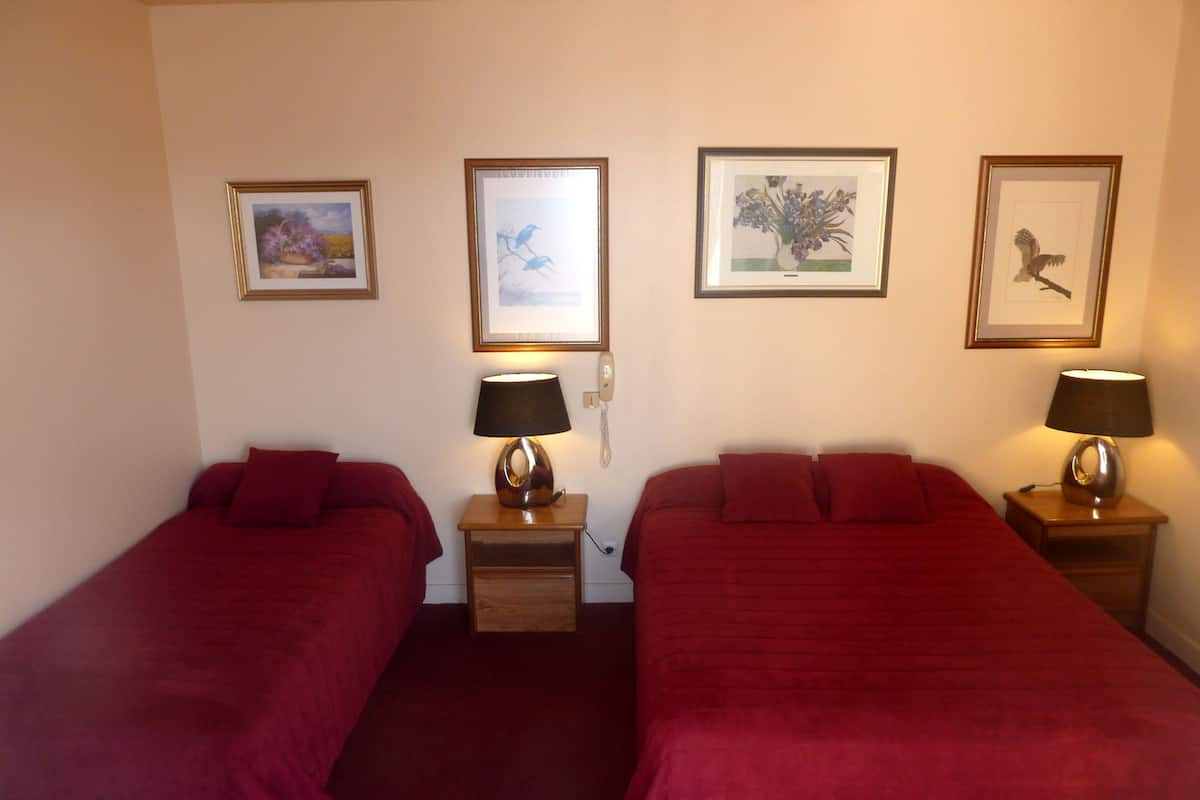 A hotel room with two single beds, each with a red bedspread and a bedside table with a lamp. There are framed pictures on the wall above each bed.