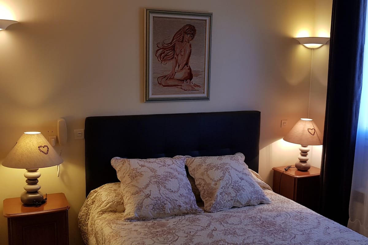 A neatly made bed with floral bedding, two bedside lamps, a framed artwork above the bed, and a window with sheer curtains.