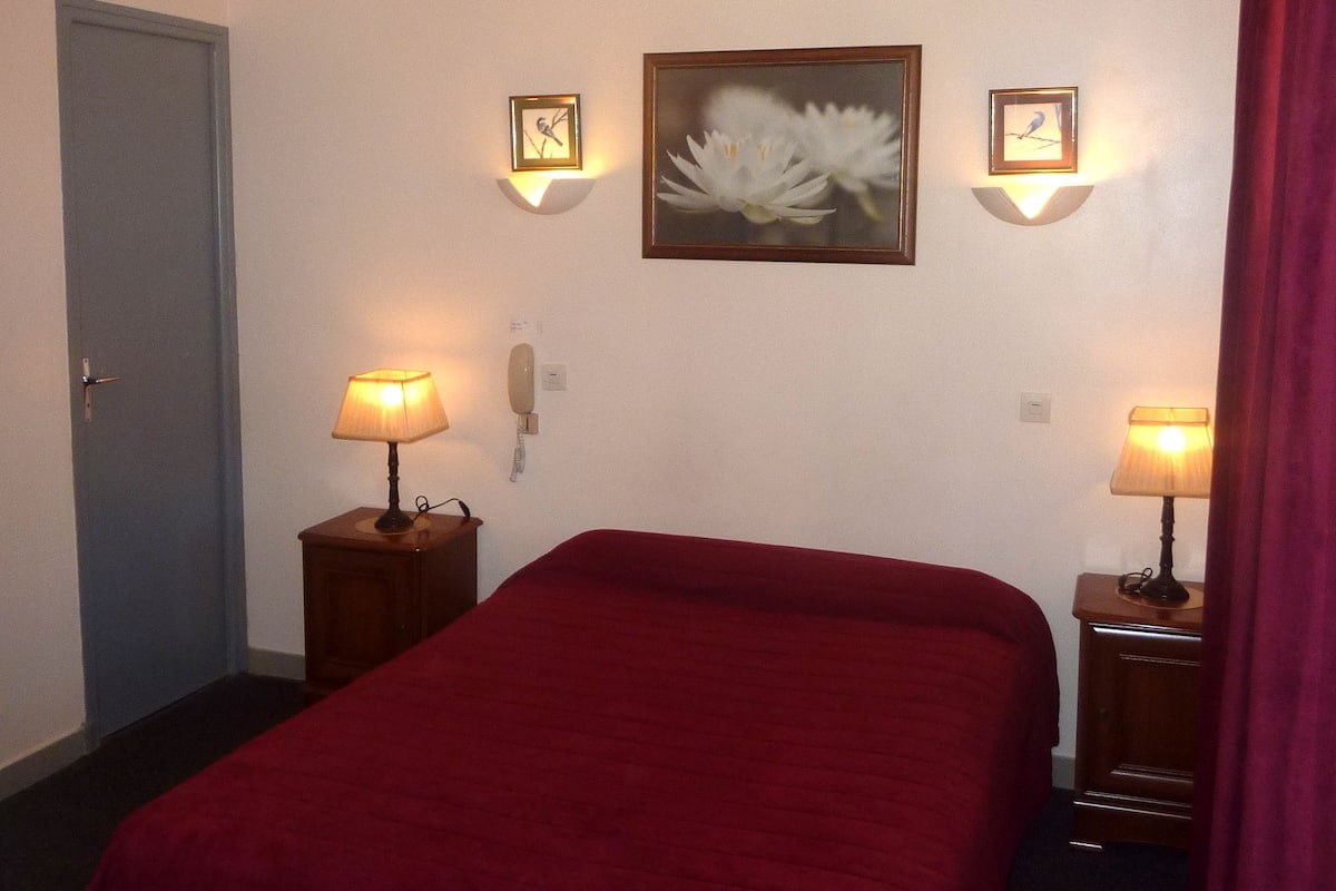 A hotel room with a single bed, two bedside tables with lamps, a wall-mounted phone, and two framed pictures.