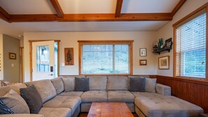 A large L-shaped sectional sofa with a wooden coffee table in the center, wooden beams on the ceiling, and large windows with blinds. - Cozy Cabin Getaway. Fireplace, king bed, hot tub, Pet OK. (Victoria)