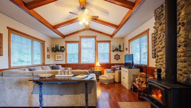 A cozy living room with a stone fireplace, wooden beams, and a large window. - Cozy Cabin Getaway. Fireplace, king bed, hot tub, Pet OK. (Victoria)