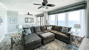 Ein großzügiges Wohnzimmer mit einem Ecksofa, einem Deckenventilator und großen Fenstern mit Blick aufs Meer.