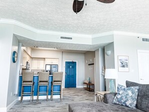 A spacious living area with a kitchen in the background, a blue door, and a grey sofa with patterned cushions.