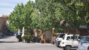 A parking lot with several parked cars, including a white SUV and a silver car. There are trees providing shade, and a building with a tiled roof is visible in the background. - A Pasturella (Monticello)